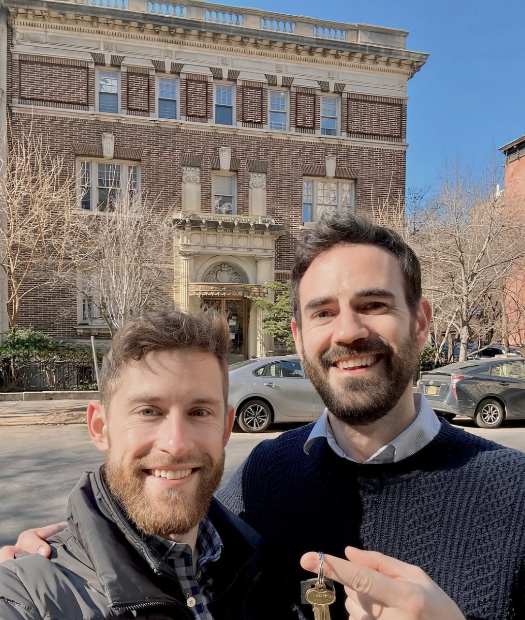 Two smiling friends holding keys in front of a brick apartment building